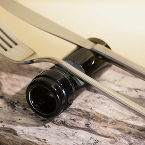 Può includere: Una forchetta e un coltello d'argento sono posizionati sopra un tappo di bottiglia nero. Il tappo di bottiglia poggia su una superficie testurizzata marrone chiaro e grigia.