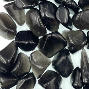May include: A collection of tumbled black obsidian stones. The stones are smooth and polished, with a dark, glassy appearance. The stones are arranged in a random pattern, with some overlapping.