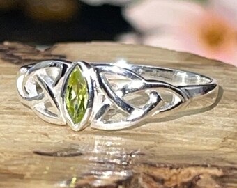 May include: A silver ring with a marquise-cut, light green gemstone. The ring features a Celtic knot design on the band. The ring is displayed on a piece of wood, with a blurred floral background.