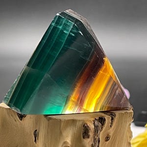 Op de afbeelding: Een groene, gele en paarse fluorietkristalpiramide op een houten basis. De kristal is gepolijst en heeft een glad oppervlak.