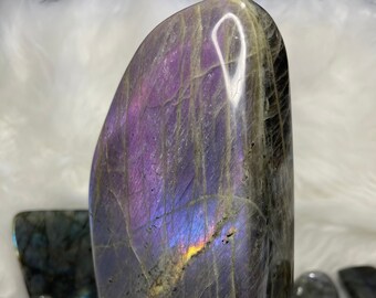 May include: A large, freeform, polished labradorite crystal with iridescent purple, blue, and green hues. The crystal is on a white surface.