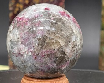 May include: A clear crystal sphere with pink and gray inclusions, resting on a wooden stand. The sphere is approximately 2 inches in diameter. The number "1" is visible on a wooden block in the foreground.