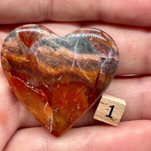 May include: A heart-shaped polished stone with red, orange, brown, and gray swirling patterns. The stone is about 2 inches wide and is sitting on a hand.