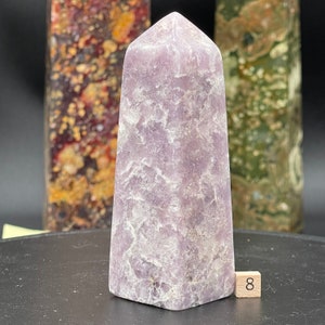 Op de afbeelding: Een lavendelkleurige kristallen obelisk met een textuur. De obelisk heeft een vierkante basis en een puntige top. Een klein houten blokje met het nummer 8 staat aan de basis. Andere decoratieve stenen obelisken staan op de achtergrond.