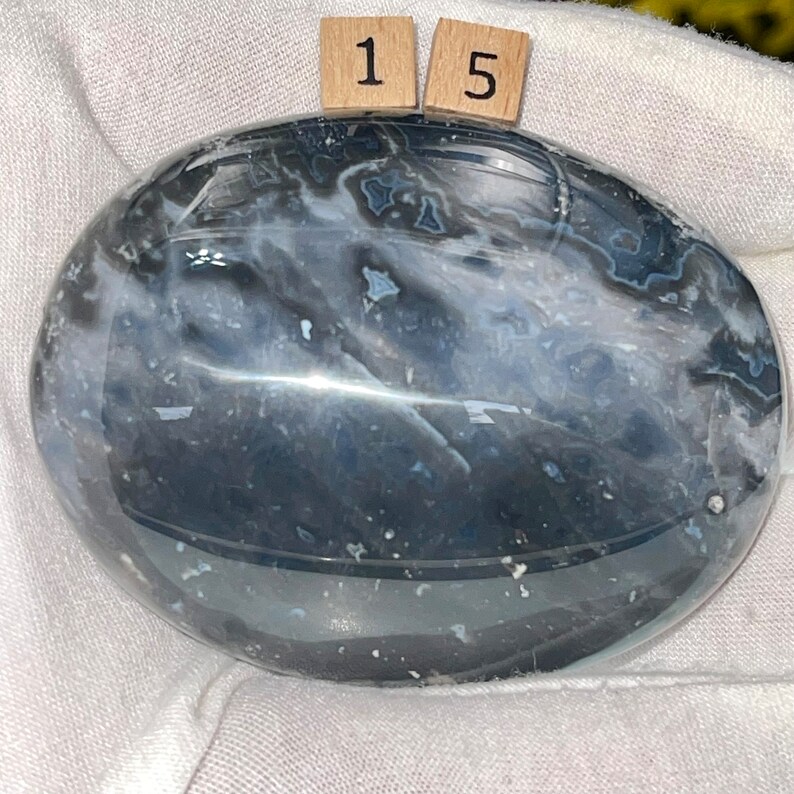 May include: A smooth, polished, oval-shaped, gray and white agate stone with a swirling pattern. The stone is approximately 2 inches in diameter.