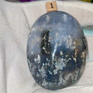 May include: A smooth, oval-shaped, blue agate stone with white and black markings. The stone is polished and has a translucent appearance.