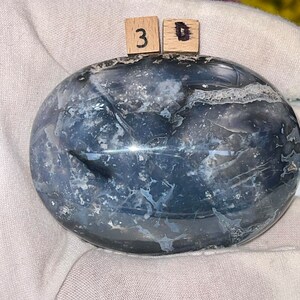 K&ouml;nnte beinhalten: Ein glatter, ovaler, polierter Achat mit einem wirbelnden Muster aus Grau, Wei&szlig; und Schwarz. Der Stein hat eine gl&auml;nzende Oberfl&auml;che und ist ein nat&uuml;rliches, einzigartiges St&uuml;ck.