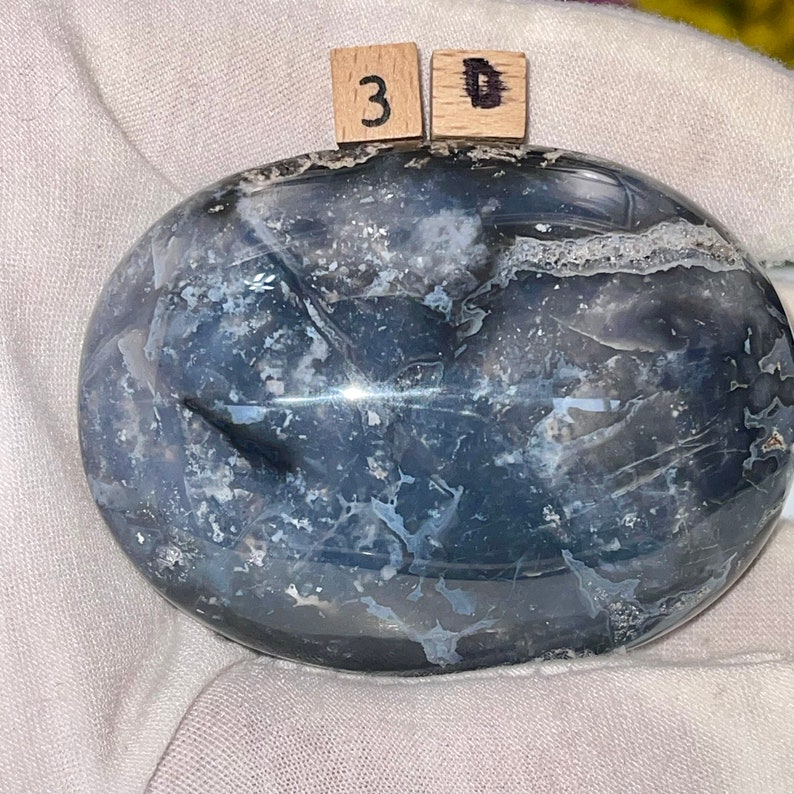 K&ouml;nnte beinhalten: Ein glatter, ovaler, polierter Achat mit einem wirbelnden Muster aus Grau, Wei&szlig; und Schwarz. Der Stein hat eine gl&auml;nzende Oberfl&auml;che und ist ein nat&uuml;rliches, einzigartiges St&uuml;ck.