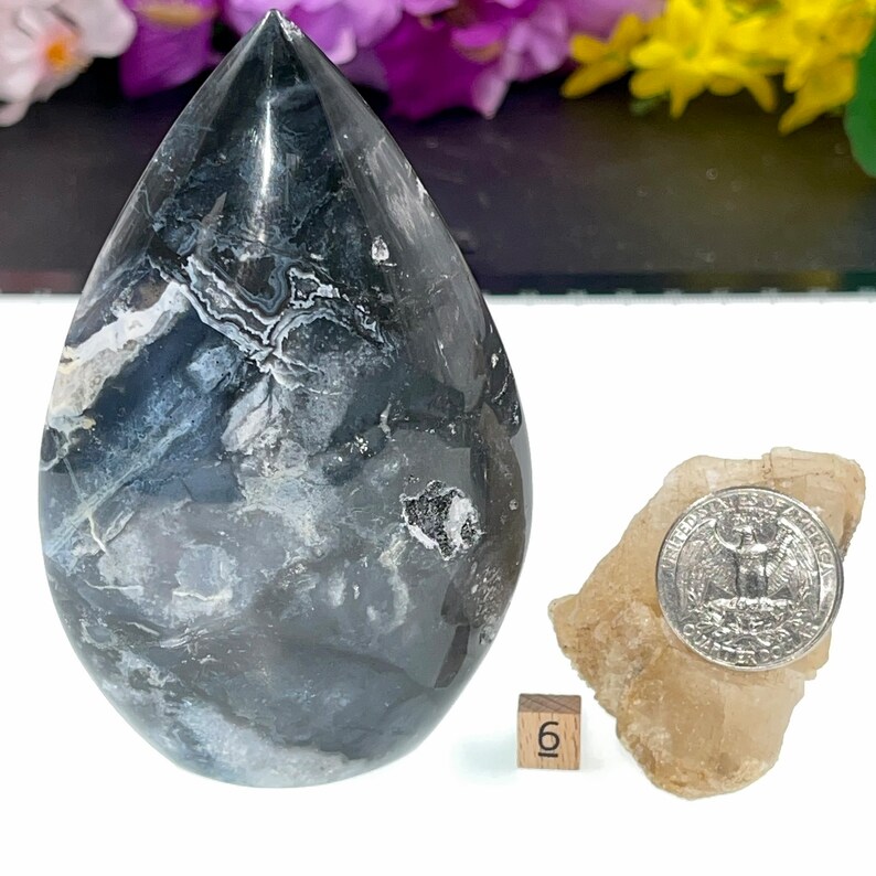May include: A teardrop-shaped black and white agate crystal with a polished finish. The crystal is approximately 6 inches tall and is sitting on a white surface. A US quarter is shown for scale.