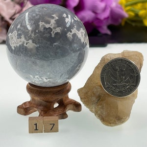 May include: A grey and white agate sphere on a wooden stand with a US quarter for scale. The sphere is approximately 1.7 inches in diameter. The sphere is sitting on a white surface with two wooden blocks labelled "1" and "7".