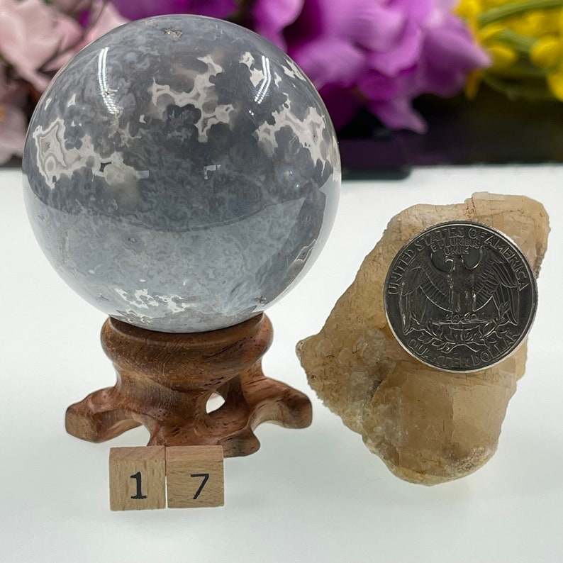 May include: A grey and white agate sphere on a wooden stand with a US quarter for scale. The sphere is approximately 1.7 inches in diameter. The sphere is sitting on a white surface with two wooden blocks labelled "1" and "7".