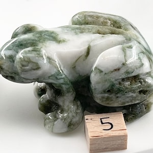 Puede incluir: Escultura detallada de una rana, hecha de piedra blanca y verde. La rana está sentada, con las patas recogidas. La piedra tiene un aspecto veteado, con vetas verdes sobre una base blanca. Un pequeño bloque de madera con el número 5 está en primer plano.