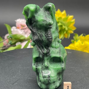 May include: A carved green and black skull figurine with a dog-like figure on top. The skull has detailed features and is made of a polished stone. The figurine is displayed on a dark surface with flowers in the background. A small wooden cube with the number 1 is in the foreground.