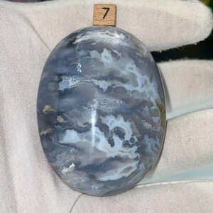 May include: A smooth, oval-shaped, polished stone with a swirling pattern of white and gray agate. The stone is labeled with the number "7".