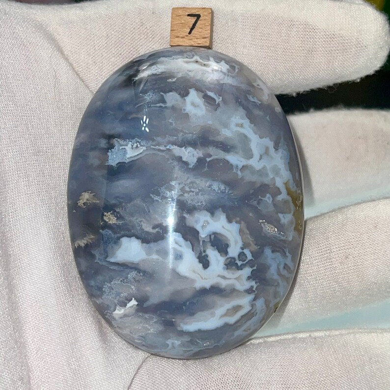 May include: A smooth, oval-shaped, polished stone with a swirling pattern of white and gray agate. The stone is labeled with the number "7".