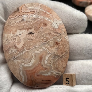 Könnte beinhalten: Ein großer, runder, polierter Achat-Stein mit einem braunen, weißen und rosafarbenen Farbmuster. Der Stein hat einen Durchmesser von etwa 5 cm.