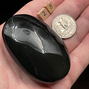 May include: A smooth, black obsidian stone, polished to a high shine, with a rounded shape. The stone is about the size of a 25 pence coin.