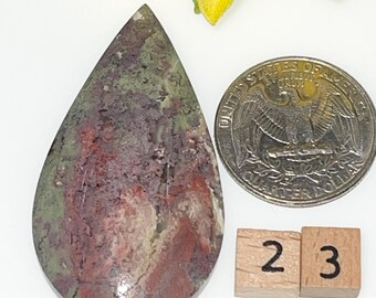 May include: A teardrop-shaped cabochon of green, red, and brown jasper, measuring approximately 2.3 inches in length. The stone is displayed next to a US quarter for size comparison.