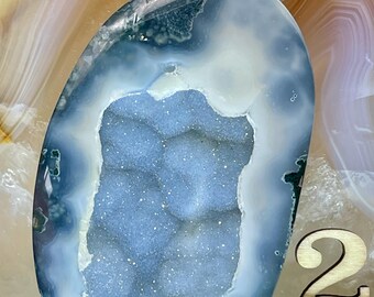 May include: A blue and white agate geode with a crystal-lined interior. The geode is shaped like a heart and has a smooth, polished surface. The geode is labeled with the number "2".
