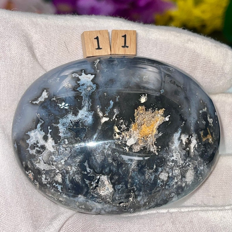 May include: A polished, oval-shaped, black and white agate stone with a light blue hue and gold flecks. The stone is labeled "11".