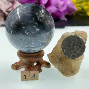 May include: A grey and white agate sphere on a wooden stand with a 25 pence coin for scale. The sphere is approximately 4 cm in diameter. The sphere is sitting on a white surface. The number 14 is written on two wooden blocks in front of the sphere.