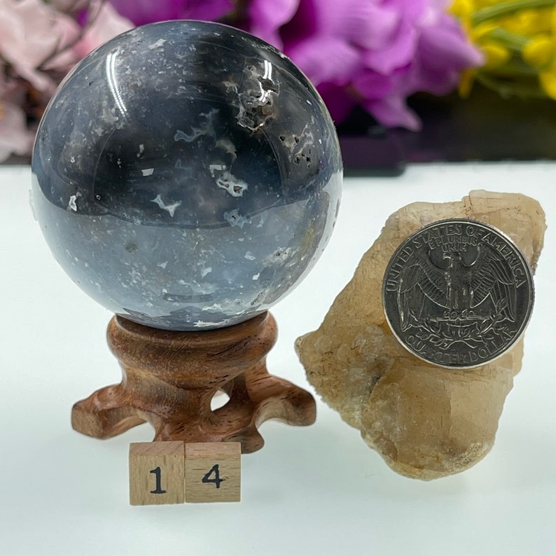 May include: A grey and white agate sphere on a wooden stand with a 25 pence coin for scale. The sphere is approximately 4 cm in diameter. The sphere is sitting on a white surface. The number 14 is written on two wooden blocks in front of the sphere.