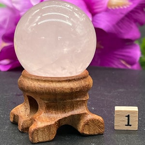 Puede incluir: Una esfera de cuarzo rosa transparente de color rosa pálido sobre un soporte de madera. La esfera tiene un diámetro de aproximadamente 5 cm. El soporte es marrón y tiene tres patas.