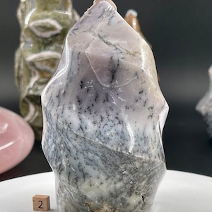 May include: A large, white, and purple, freeform, polished, crystal point with black dendritic inclusions. The crystal is sitting on a white surface with a small wooden block with the number "2" on it.