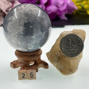 May include: A grey and white agate sphere on a wooden stand with a 26 label. The sphere is approximately 5 cm in diameter. A small piece of raw selenite is in the background.