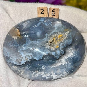 K&ouml;nnte beinhalten: Ein glatter, ovaler blauer Achat-Stein mit wei&szlig;en und goldenen Markierungen. Der Stein ist poliert und hat ein nat&uuml;rliches, organisches Aussehen. Die Zahlen "26" sind auf zwei kleinen Holzbl&ouml;cken geschrieben.