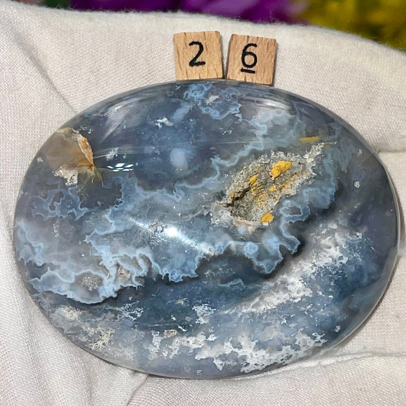 K&ouml;nnte beinhalten: Ein glatter, ovaler blauer Achat-Stein mit wei&szlig;en und goldenen Markierungen. Der Stein ist poliert und hat ein nat&uuml;rliches, organisches Aussehen. Die Zahlen "26" sind auf zwei kleinen Holzbl&ouml;cken geschrieben.