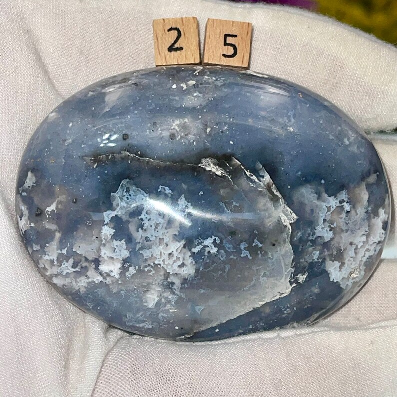 K&ouml;nnte beinhalten: Ein glatter, ovaler blauer Achat-Stein mit wei&szlig;en und grauen Markierungen. Der Stein ist poliert und hat eine gl&auml;nzende Oberfl&auml;che. Die Zahlen "25" sind auf zwei kleinen Holzbl&ouml;cken geschrieben.