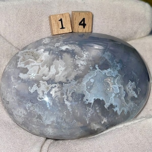 May include: A smooth, gray and white agate stone with a cloudy pattern. The stone is polished and has a natural, organic shape. The stone is labeled with the number "14".