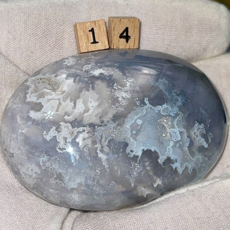 May include: A smooth, gray and white agate stone with a cloudy pattern. The stone is polished and has a natural, organic shape. The stone is labeled with the number "14".