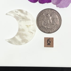 May include: White crescent moon shaped crystal with a cloudy pattern. The crystal is about the size of a US quarter. A wooden tile with the number 6 is in the background.