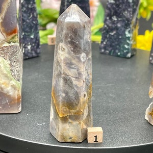 Op de afbeelding: Een grijze, witte en bruine agaatpunt met een gepolijste afwerking. De punt is ongeveer 5 cm hoog en heeft een natuurlijk, organisch patroon. De punt ligt op een zwart oppervlak met een klein houten blokje met het opschrift "1".