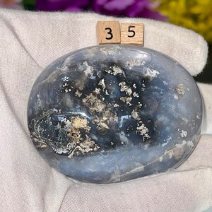 May include: A smooth, gray and white agate stone with a unique pattern of black and silver inclusions. The stone is polished and has a rounded shape. The numbers "35" are written on two small wooden blocks.