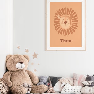 K&ouml;nnte beinhalten: Ein gerahmter Druck mit einem pfirsichfarbenen Hintergrund, der einen Comic-L&ouml;wen mit einer braunen M&auml;hne und dem Namen "Theo" darunter zeigt. Der Druck h&auml;ngt an einer wei&szlig;en Wand &uuml;ber einem Bett mit einem gro&szlig;en Teddyb&auml;r und anderen Stofftieren.