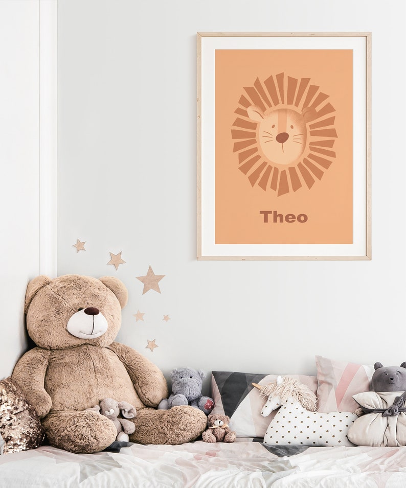 K&ouml;nnte beinhalten: Ein gerahmter Druck mit einem pfirsichfarbenen Hintergrund, der einen Comic-L&ouml;wen mit einer braunen M&auml;hne und dem Namen "Theo" darunter zeigt. Der Druck h&auml;ngt an einer wei&szlig;en Wand &uuml;ber einem Bett mit einem gro&szlig;en Teddyb&auml;r und anderen Stofftieren.