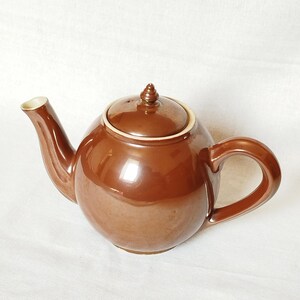 May include: A brown ceramic teapot with a rounded body, a curved spout, and a loop handle. The lid has a small knob on top. The rim of the spout and the lid are trimmed with a cream-colored band. The teapot is set against a white background.