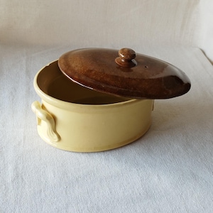 May include: A ceramic casserole dish with a lid. The dish is light yellow with two side handles. The lid is brown with a small knob. The lid is slightly ajar, revealing the interior of the dish.