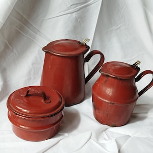 Puede incluir: Tres recipientes de cocina de esmalte rojo con tapas. El recipiente más grande tiene un pico y un asa. El recipiente más pequeño tiene una tapa con un asa.
