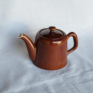 Könnte beinhalten: Kleine, braune Keramik-Teekanne mit Deckel und Henkel. Die Teekanne hat einen gebogenen Ausguss und einen kleinen Knopf auf dem Deckel. Die Teekanne steht auf einer weißen Oberfläche.