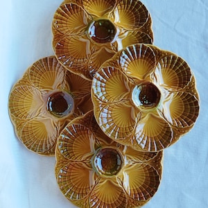 Pode incluir: Quatro pratos de ostras de cerâmica numa paleta de cores quentes e terrosas. Cada prato apresenta um design em forma de concha com compartimentos individuais e um poço central. Os pratos estão dispostos sobre uma superfície branca.