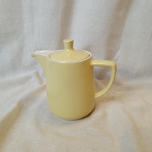 Puede incluir: Una jarra de crema de cerámica de color amarillo pálido con una tapa blanca y un asa. La jarra de crema está sobre un fondo de tela blanca.