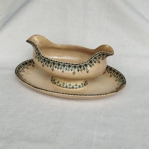 Puede incluir: Un juego de salsera y platillo de color crema. El recipiente tiene una forma curva con un pico a cada lado. El borde y el borde del platillo están decorados con un estampado floral verde oscuro. El juego es de cerámica.