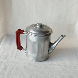 Puede incluir: Una cafetera vintage de aluminio con un asa roja. La cafetera tiene una tapa y un pico para verter. El asa es de color rojo brillante, contrastando con el plateado de la cafetera. La cafetera es probablemente de mediados del siglo XX.