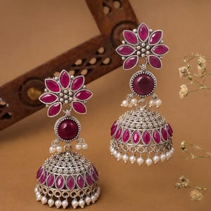 May include: A pair of silver-toned earrings with a floral design. Each earring features a flower-shaped top with maroon-colored petals, a round maroon stone, and a bell-shaped bottom adorned with maroon stones and small white pearls. The earrings are displayed against a brown background.