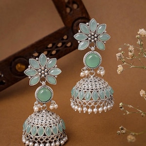 May include: A pair of silver-toned earrings with a floral design. Each earring features a flower-shaped top, a round green stone, and a bell-shaped bottom adorned with green stones and small white pearls. The earrings have an antique finish.