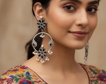 May include: Silver-toned earrings with black floral and teardrop accents, adorned with small white pearls. The earrings feature a circular design and are worn with a colorful embroidered top.